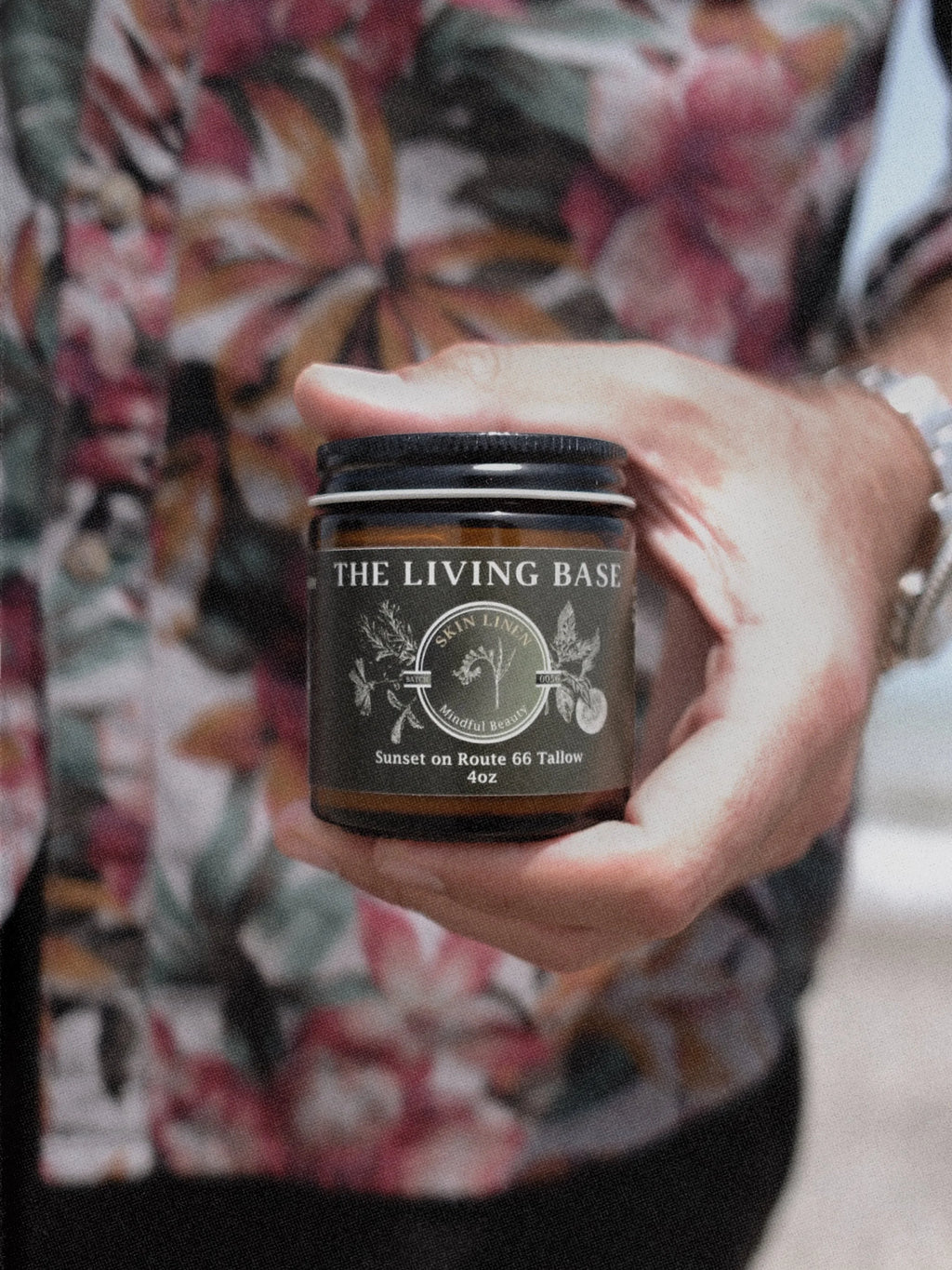Person holding a jar of 'The Living Base' product with a floral shirt in the background